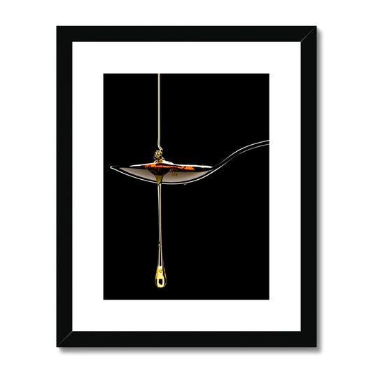 Honey pouring on to metal spoon and dripping off against black background. Framed & Mounted Print