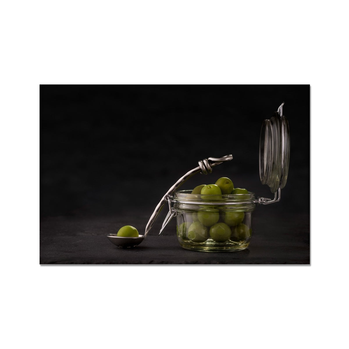 Jar of green olives with single olive on spoon against grey slate background. Fine Art Print