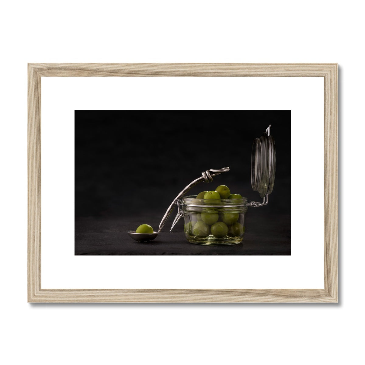 Jar of green olives with single olive on spoon against grey slate background. Framed & Mounted Print