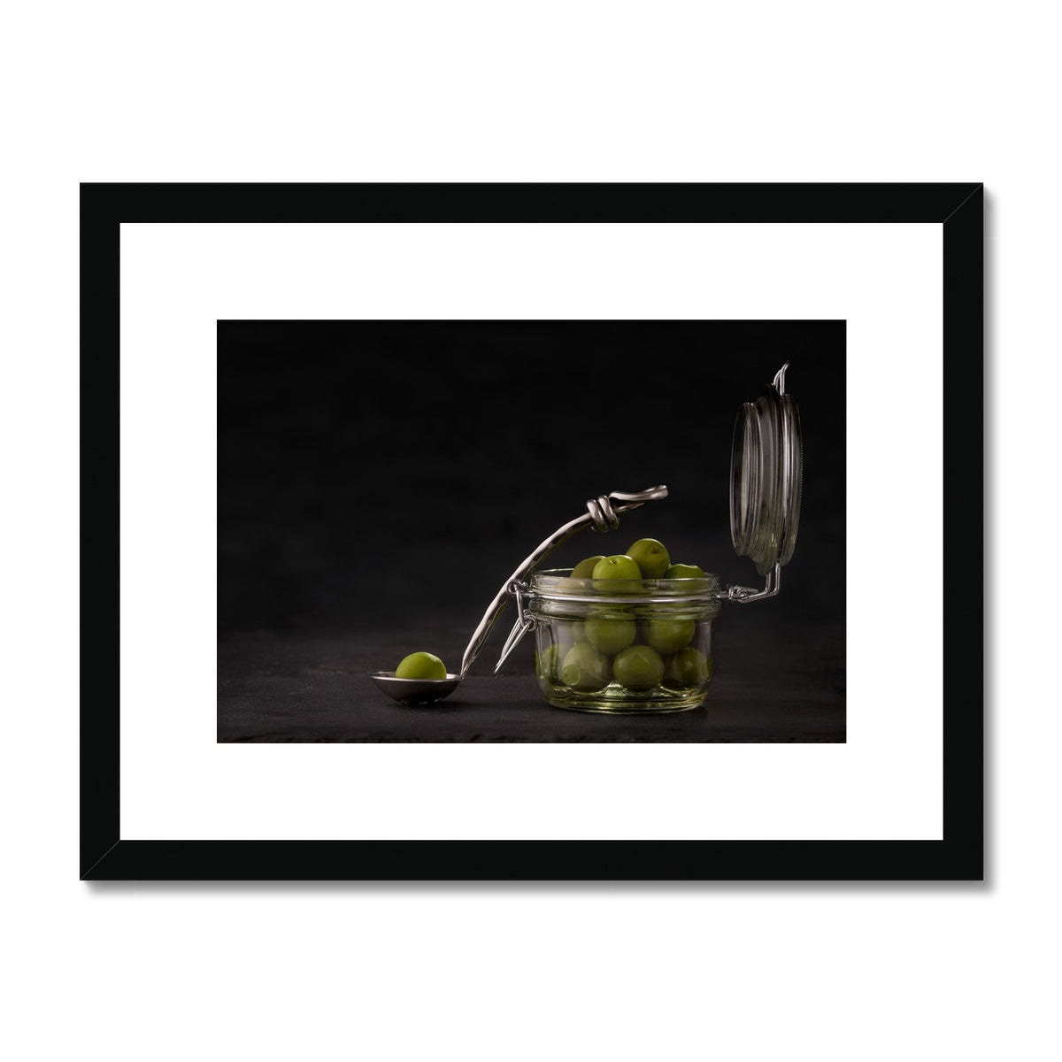 Jar of green olives with single olive on spoon against grey slate background. Framed & Mounted Print