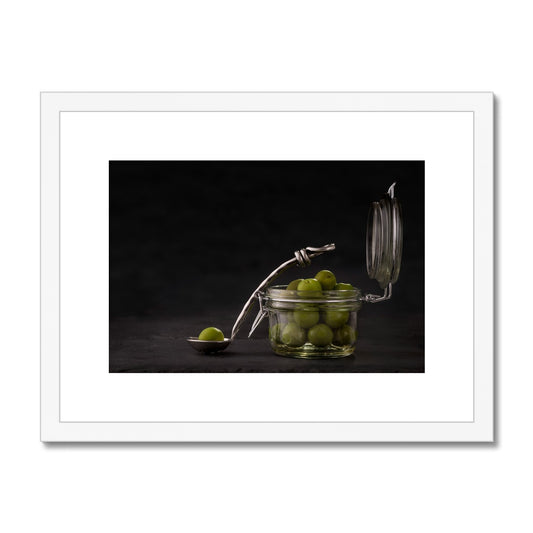 Jar of green olives with single olive on spoon against grey slate background. Framed & Mounted Print